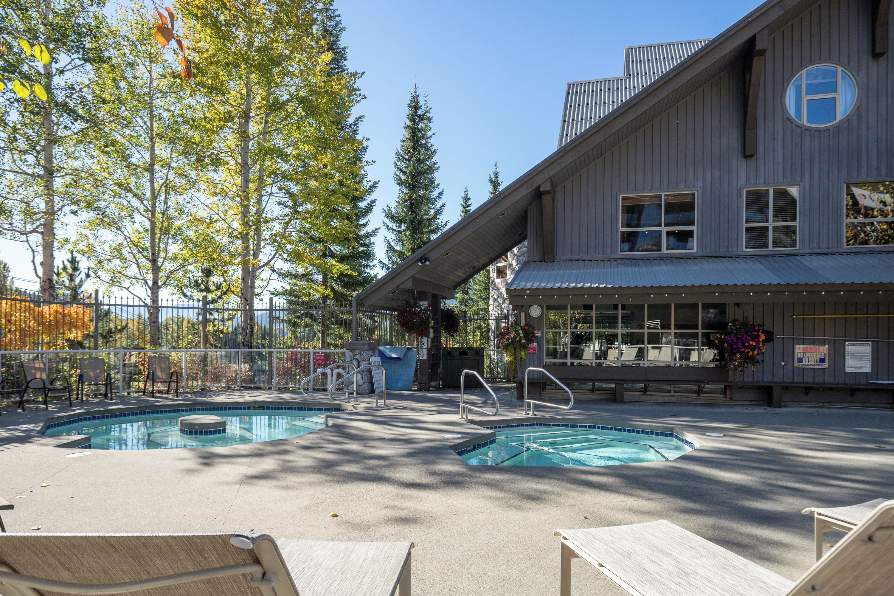 A pool area with lounge chairs, a building with a large window, and a fence with trees.