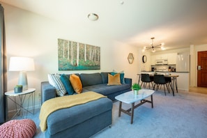 Un salon comprenant un canapé, une table basse, une table à manger avec des chaises, un coin cuisine et un tableau accroché au mur.