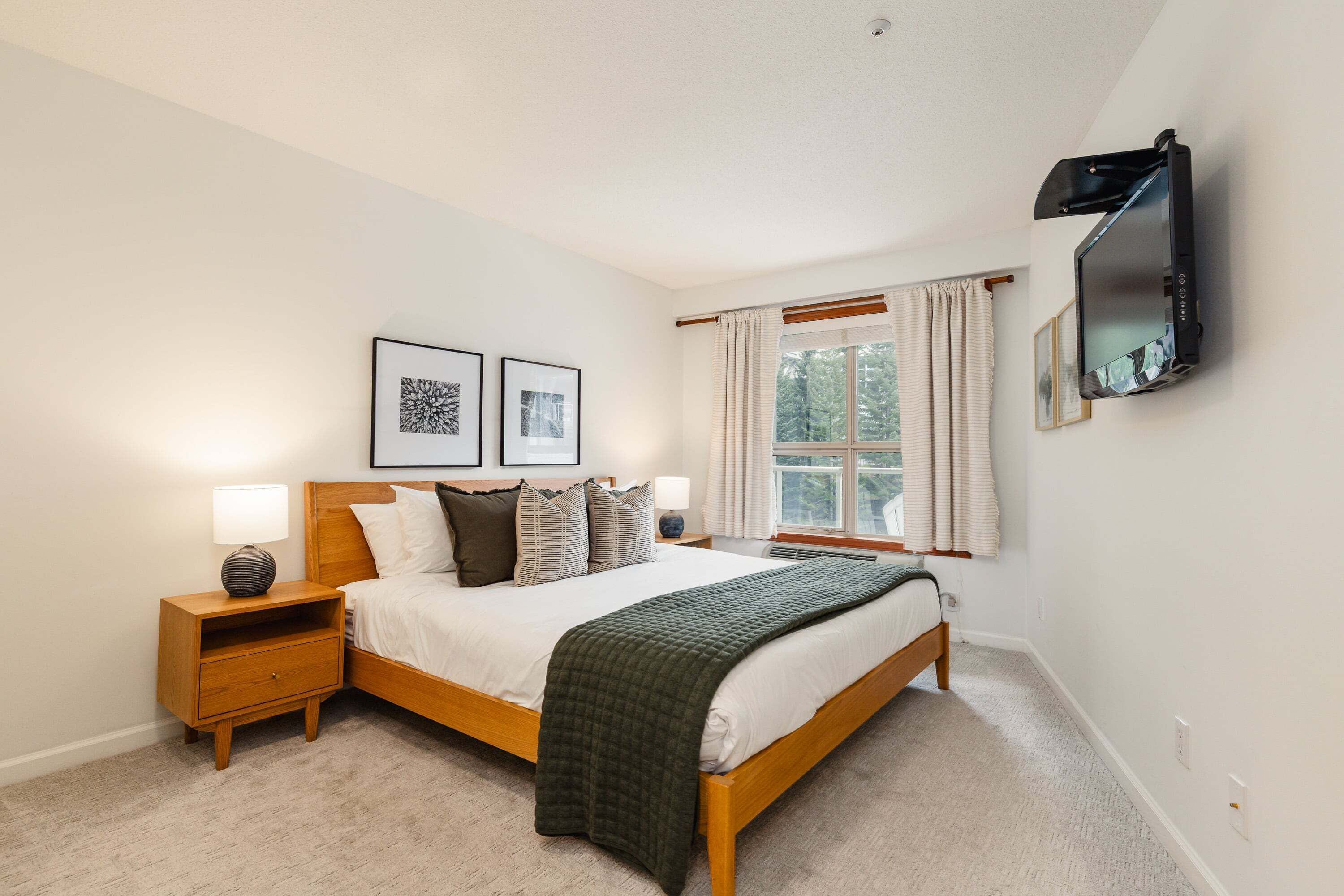 A bedroom with a bed, nightstands, a TV mounted on the wall, and a window with curtains.
