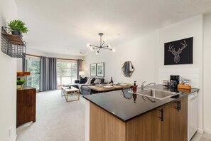 A modern kitchen with a central island, a dining area with a sofa, and a living room with a large window. - Panorama Suite at the Aspens (Whistler)