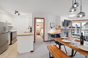 A modern kitchen with white cabinetry, a dining area with a wooden table and chairs, and a view through a large window. - Powder Peak at the Aspens (Whistler)