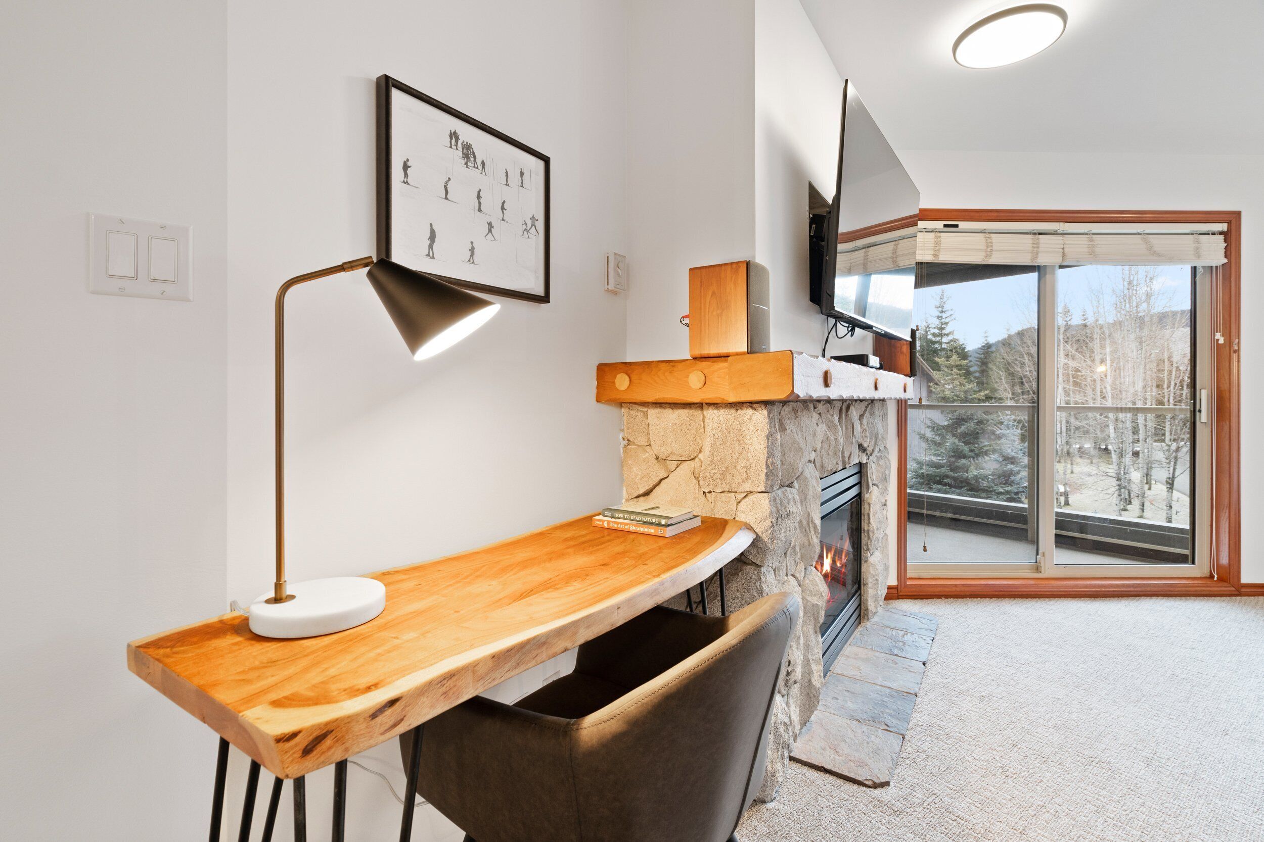 Una sala de estar moderna con chimenea, una mesa de madera, una lámpara y un ventanal con vistas a árboles.