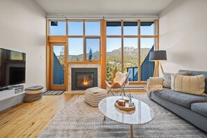 A modern living room with a fireplace, large windows, a sofa, a coffee table, and a TV. - Ridgeline Retreat at Sundance (Whistler)
