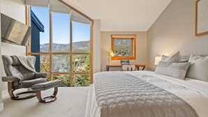 A bedroom with a bed, a reclining chair, a desk, a window with a view of the mountains, and a painting on the wall. - Ridgeline Retreat at Sundance (Whistler)