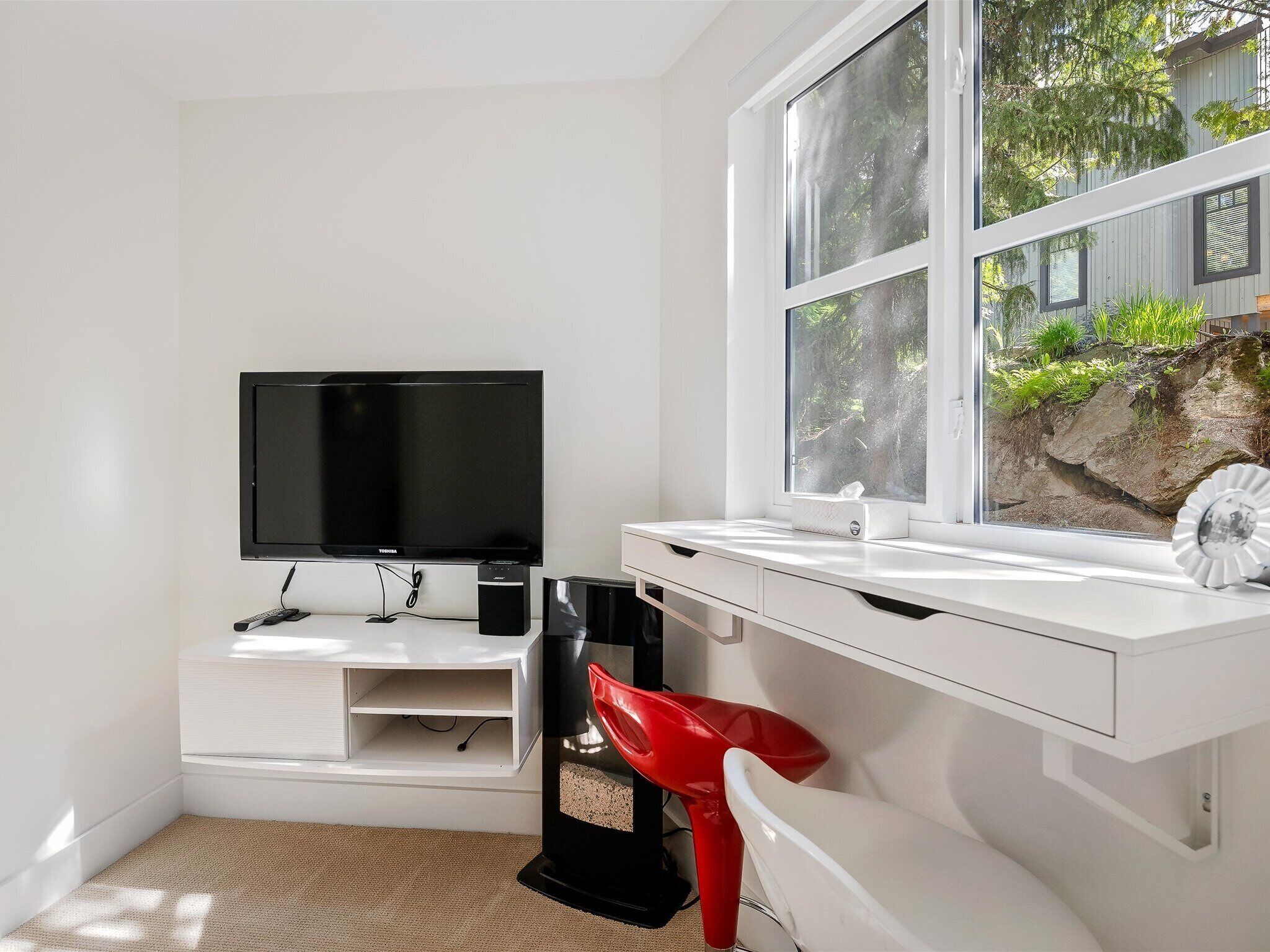 A modern, white room with a large window, a flat-screen TV, a desk, and a red chair.