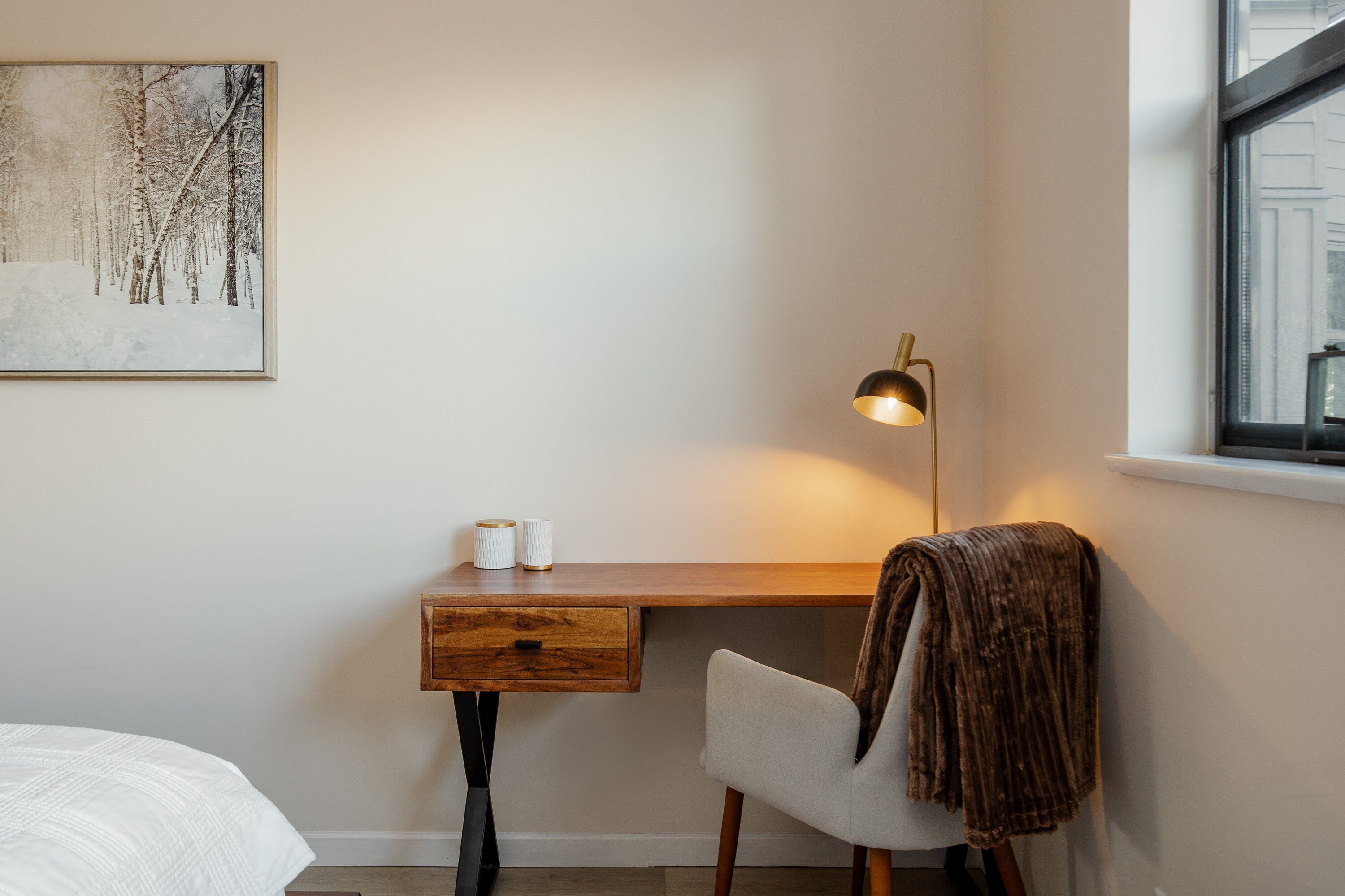 Quarto com escrivaninha de madeira, uma cadeira, mesa de cabeceira, abajur e um quadro na parede.