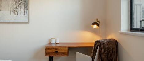 Quarto com escrivaninha de madeira, uma cadeira, mesa de cabeceira, abajur e um quadro na parede.