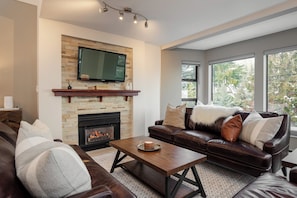 A living room with a fireplace, leather sofa, and a flat-screen TV mounted on a brick wall. - Seven Pines at Greyhawk (Whistler)