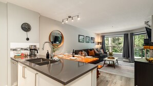 A modern kitchen with a central island, a dining area with a sofa, and a large window with curtains. - Skywalk Suite at the Aspens (Whistler)