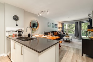 A modern kitchen with a central island, a dining area with a sofa, and a large window with curtains. - Skywalk Suite at the Aspens (Whistler)