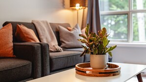 A living room with a sofa, a potted plant, a lamp, and a window with a view of trees. - Skywalk Suite at the Aspens (Whistler)
