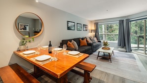 A dining area with a wooden table set for four, a round mirror, and a sofa in the background. - Skywalk Suite at the Aspens (Whistler)