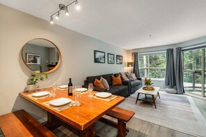 A dining area with a wooden table set for four, a round mirror, and a sofa in the background. - Skywalk Suite at the Aspens (Whistler)