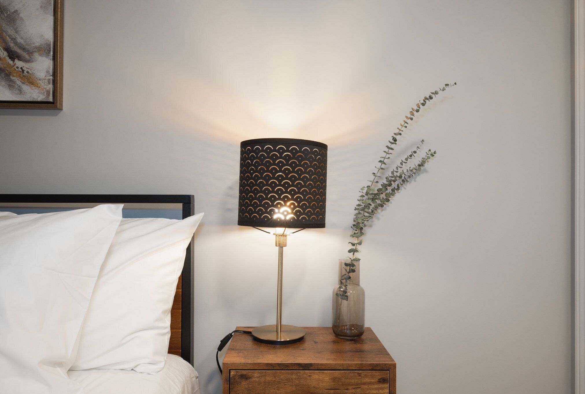 Een nachtkastje met een lamp, een vaas met een plant en een bed met wit beddengoed.