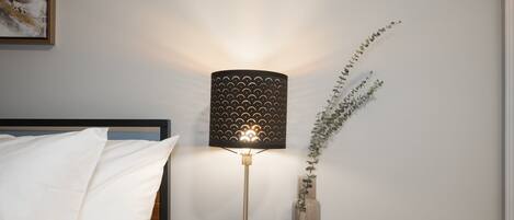 Een nachtkastje met een lamp, een vaas met een plant en een bed met wit beddengoed.