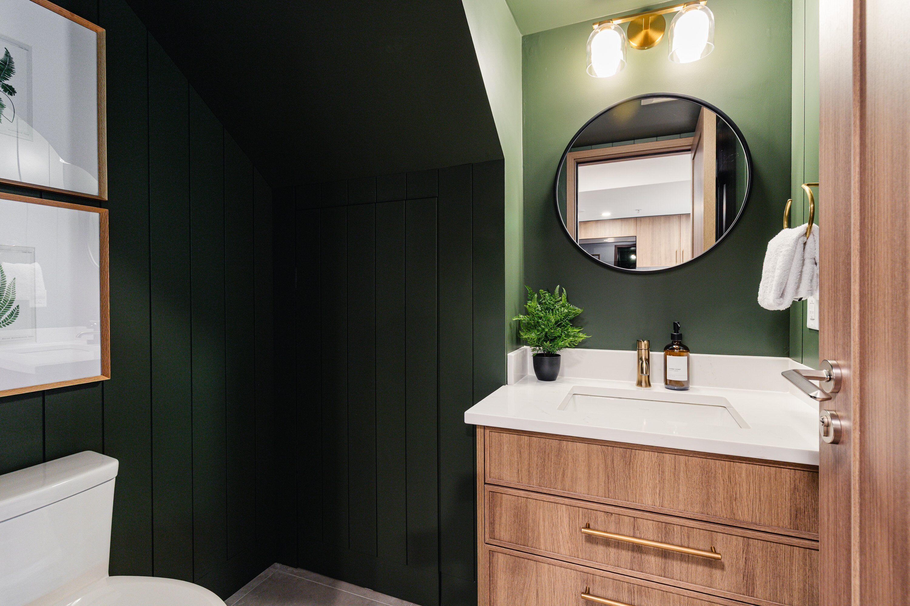 Une salle de bain avec des toilettes blanches, un meuble-lavabo en bois avec un miroir, un évier et une plante en pot.