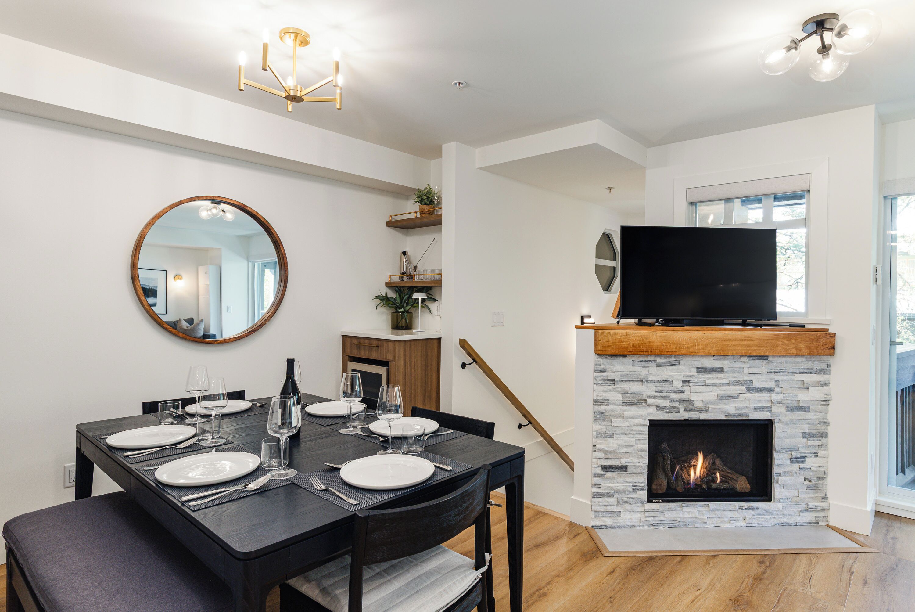 A dining area with a black table set for four, a round mirror, and a stone fireplace with a TV above it.
