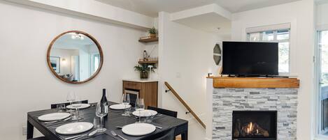 A dining area with a black table set for four, a round mirror, and a stone fireplace with a TV above it.