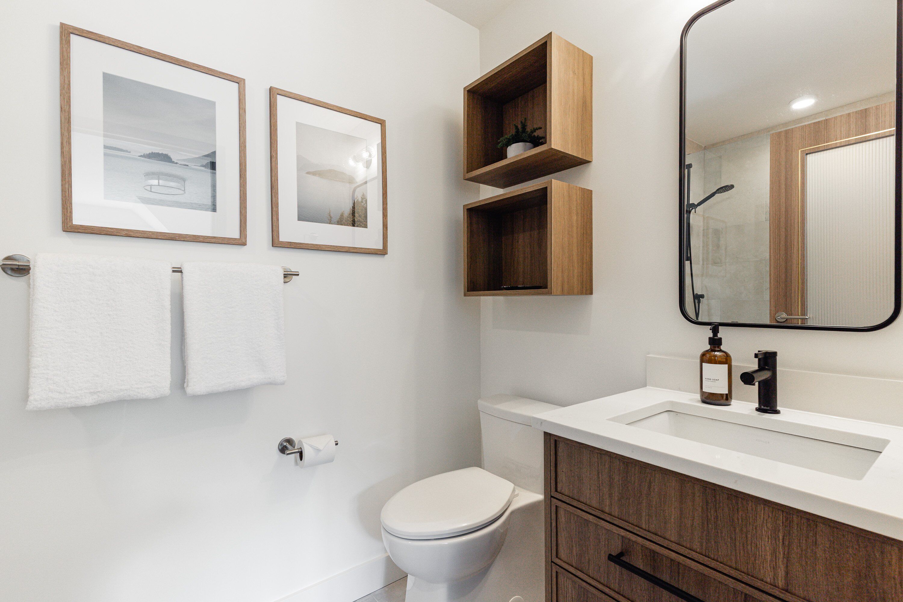 Une salle de bain avec des toilettes, un lavabo, des étagères en bois, des tableaux encadrés et un miroir.