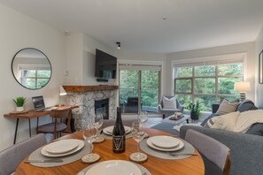 A dining area with a round table, chairs, and a bottle of wine. There is a fireplace, a television, and a window with a view of trees. - The Treehouse at the Aspens (Whistler)