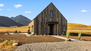 A rustic wooden cabin with a large window, surrounded by a gravel driveway and a small patio area. - Glass House Montana | The valley, without compromise (Pray)