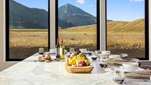 A dining table with a fruit bowl, wine bottles, and glasses, set against a backdrop of a mountain landscape through large windows. - Glass House Montana | The valley, without compromise (Pray)