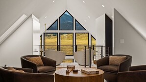 A modern interior with a high ceiling, large windows offering a view of a mountainous landscape, and a seating area with leather sofas and a wooden coffee table. - Glass House Montana | The valley, without compromise (Pray)