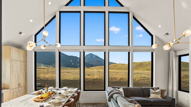 A modern interior with large windows offering a mountain view, a dining area with a table set, and a cozy seating area. - Glass House Montana | The valley, without compromise (Pray)