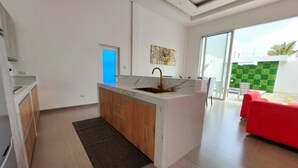 A modern kitchen with a central island, a sink, and a window. There is a red couch and a green wall in the adjacent living area.