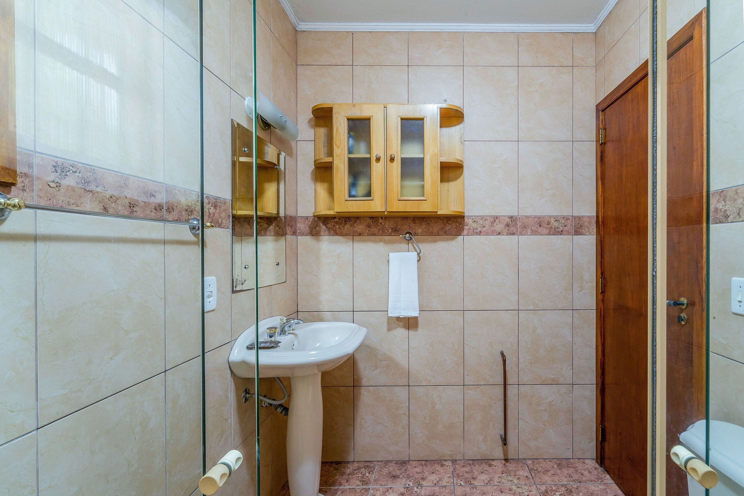 A bathroom with a glass shower enclosure, a pedestal sink, a wooden cabinet with frosted glass doors, and a marble-tiled floor.
