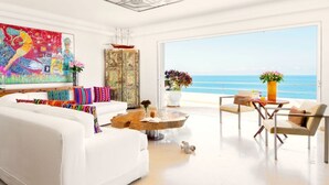A modern living room with a white sofa, colorful pillows, a round coffee table, and a view of the ocean.