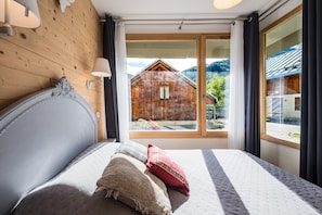 A bedroom with a large bed, a wooden headboard, a window with a view of a wooden cabin, and a lamp.
