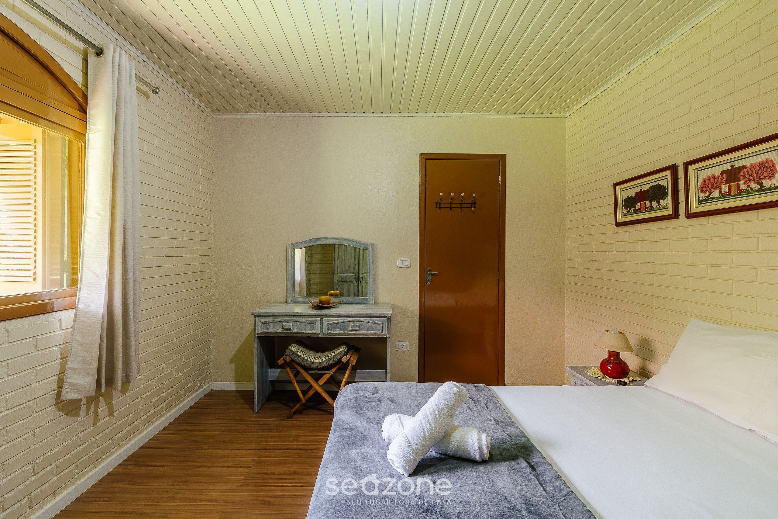 A bedroom with a bed, a wooden desk, a chair, a mirror, a door, and a nightstand with a lamp.