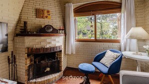 A cozy room with a brick fireplace, a blue armchair, and a window with curtains. - House With Barbecue in Gramado Rje0980 (Gramado)