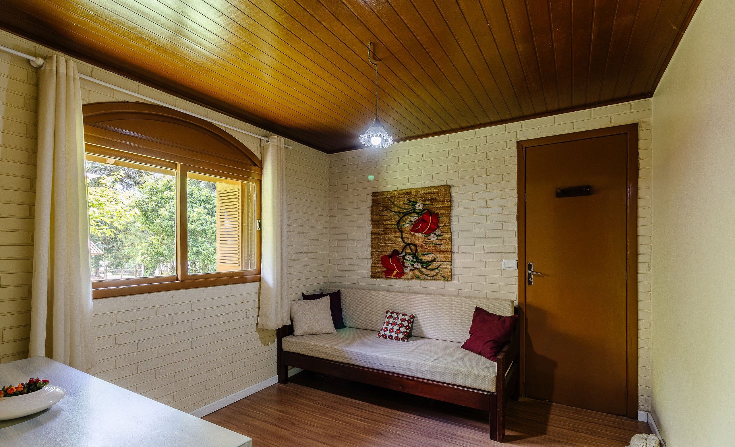 A cozy room with a wooden ceiling, a white brick wall, a wooden door, and a cushioned bench with pillows.