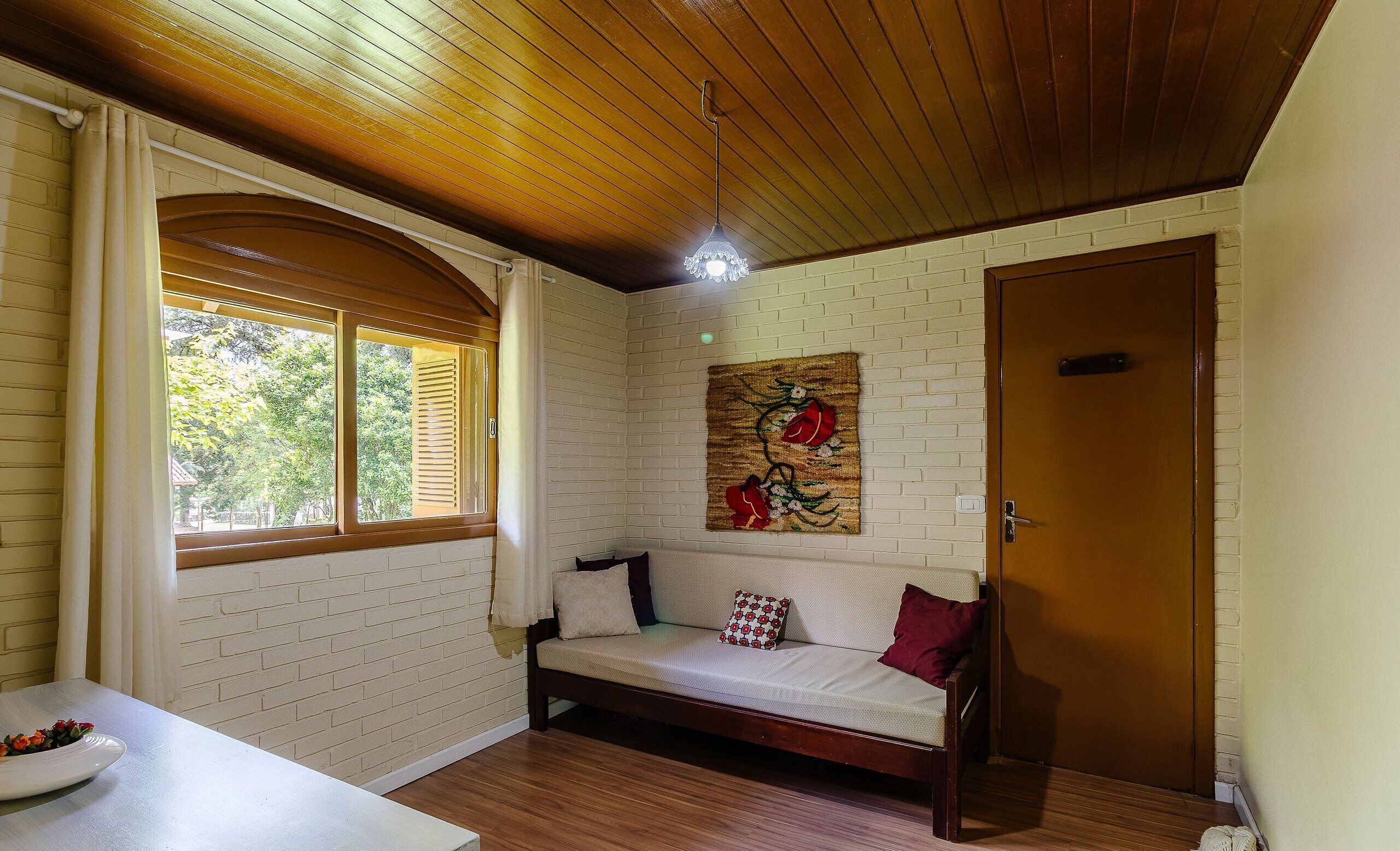 A cozy room with a wooden ceiling, a white brick wall, a wooden door, and a cushioned bench with pillows.