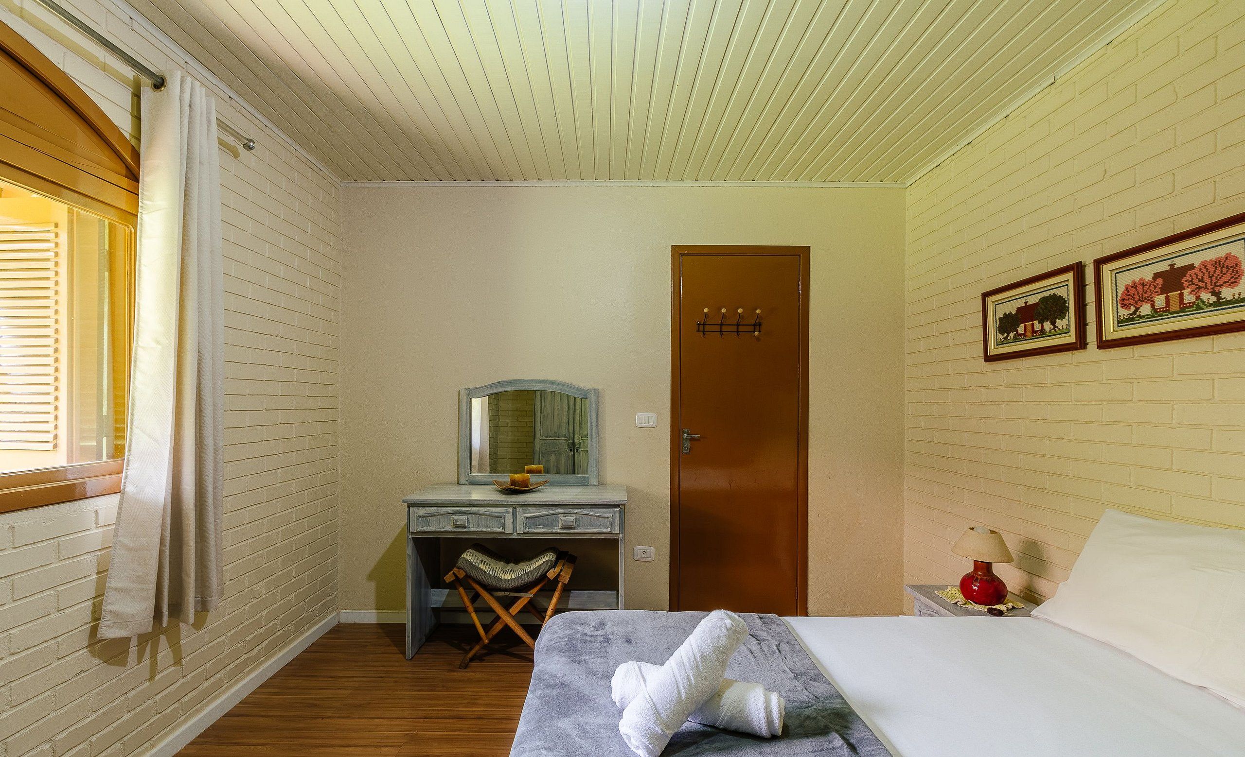 Un dormitorio con cama, un escritorio de madera, una silla, un espejo, una puerta y una mesita de noche con lámpara.