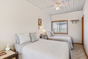 A bedroom with two beds, a ceiling fan, a window with blinds, a bedside table with a lamp, and a wall-mounted mirror.