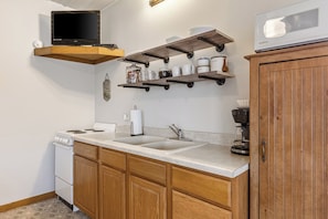 A kitchen with a stove, sink, wooden cabinets, a coffee maker, and a television mounted on a wooden shelf.