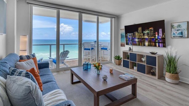 A living room with a large sliding glass door leading to a balcony with a view of the ocean and a city skyline. There is a sofa, a coffee table, and a TV mounted on the wall. - Elara Beachfront 2 BRS Direct OceanView w Parking (Miami Beach)