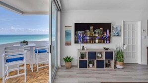 A balcony with a view of the ocean, a seating area with blue cushions, a television mounted on the wall, and a wooden console table with decorative items. - Elara Beachfront 2 BRS Direct OceanView w Parking (Miami Beach)