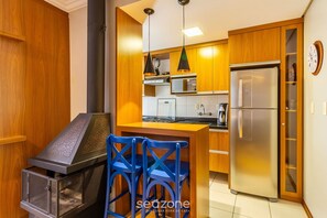 A compact kitchen with wooden cabinets, a stainless steel refrigerator, a microwave, and a built-in oven. There are blue chairs and a small bar area with a grill. - Apt w/ BBQ 1km From Gramado Center Sds104 (Gramado)