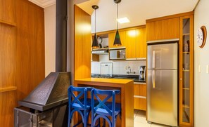 A compact kitchen with wooden cabinets, a stainless steel refrigerator, a microwave, and a built-in oven. There are blue chairs and a small bar area with a grill. - Apt w/ BBQ 1km From Gramado Center Sds104 (Gramado)