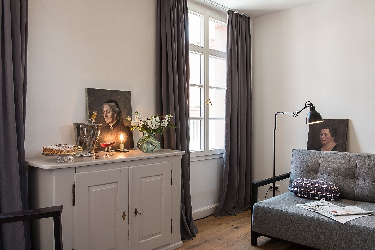 A room with a grey sofa, a white cabinet, a painting, and a lamp.