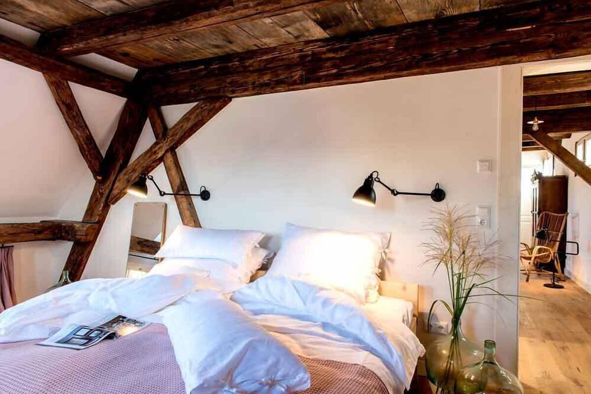 Une chambre avec un plafond en bois, un lit recouvert de draps blancs, deux lampes de chevet et un vase contenant des plantes séchées.