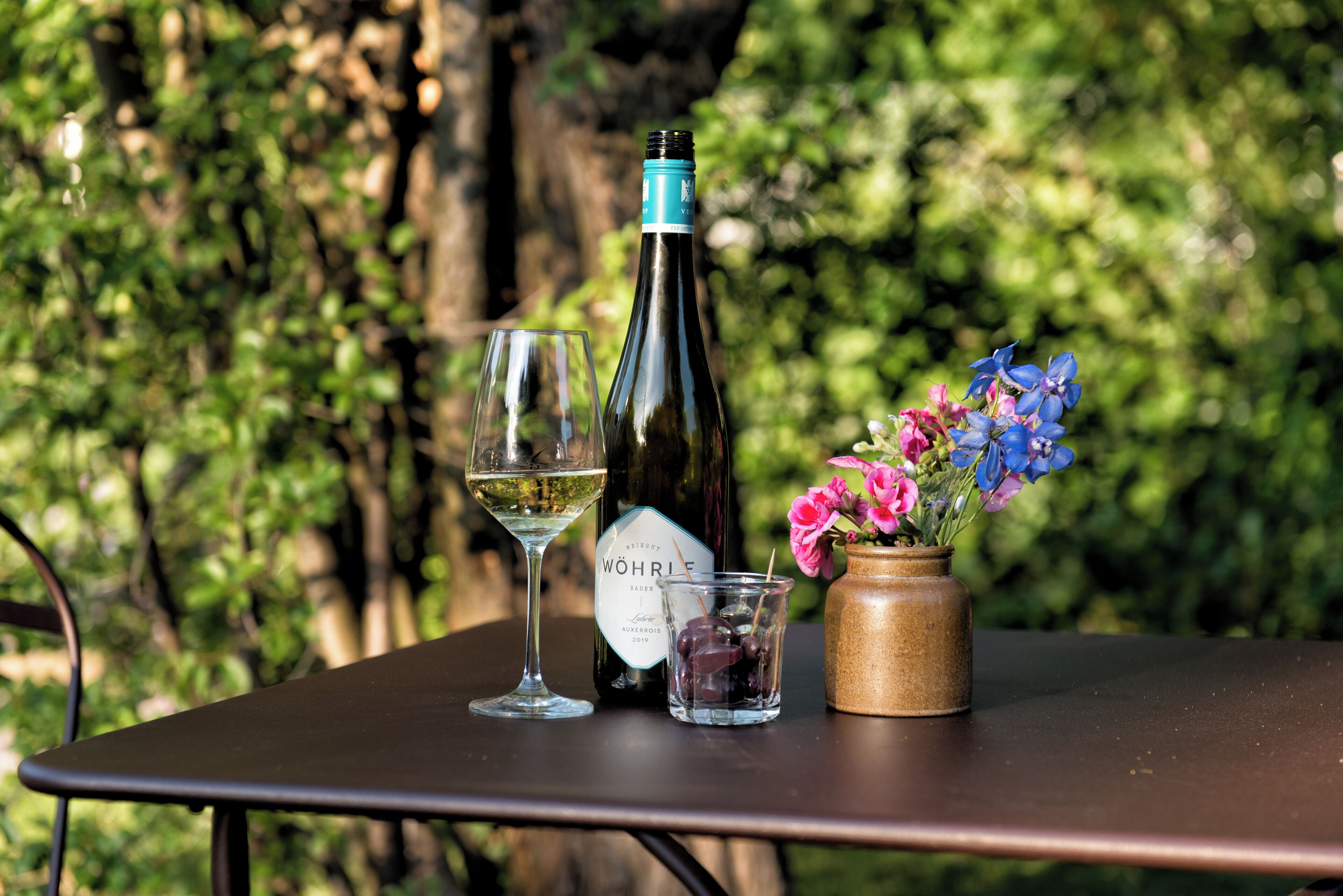 A table with a bottle of wine, a wine glass, and a small glass with olives and grapes.