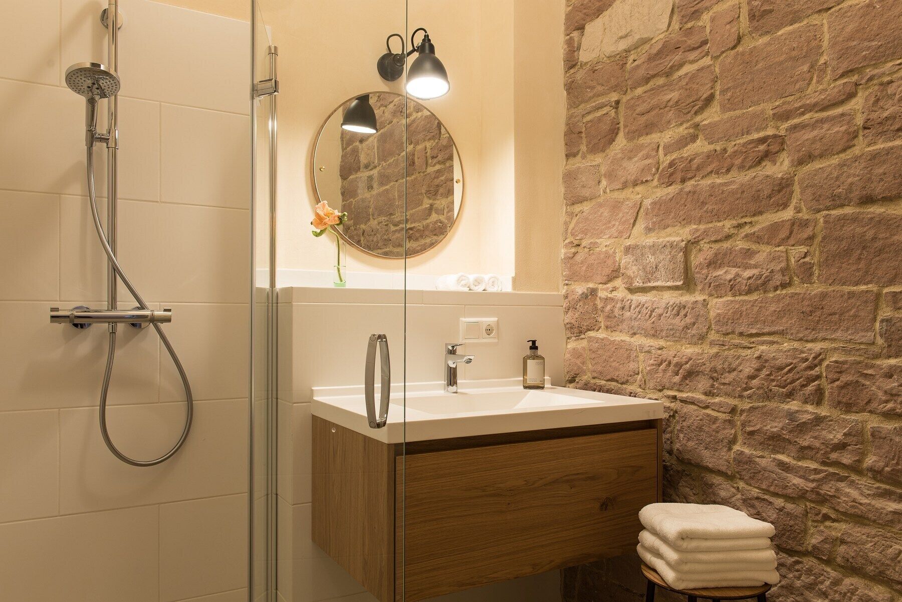 A bathroom with a glass shower enclosure, a wooden vanity with a sink, a mirror, and a stone wall.