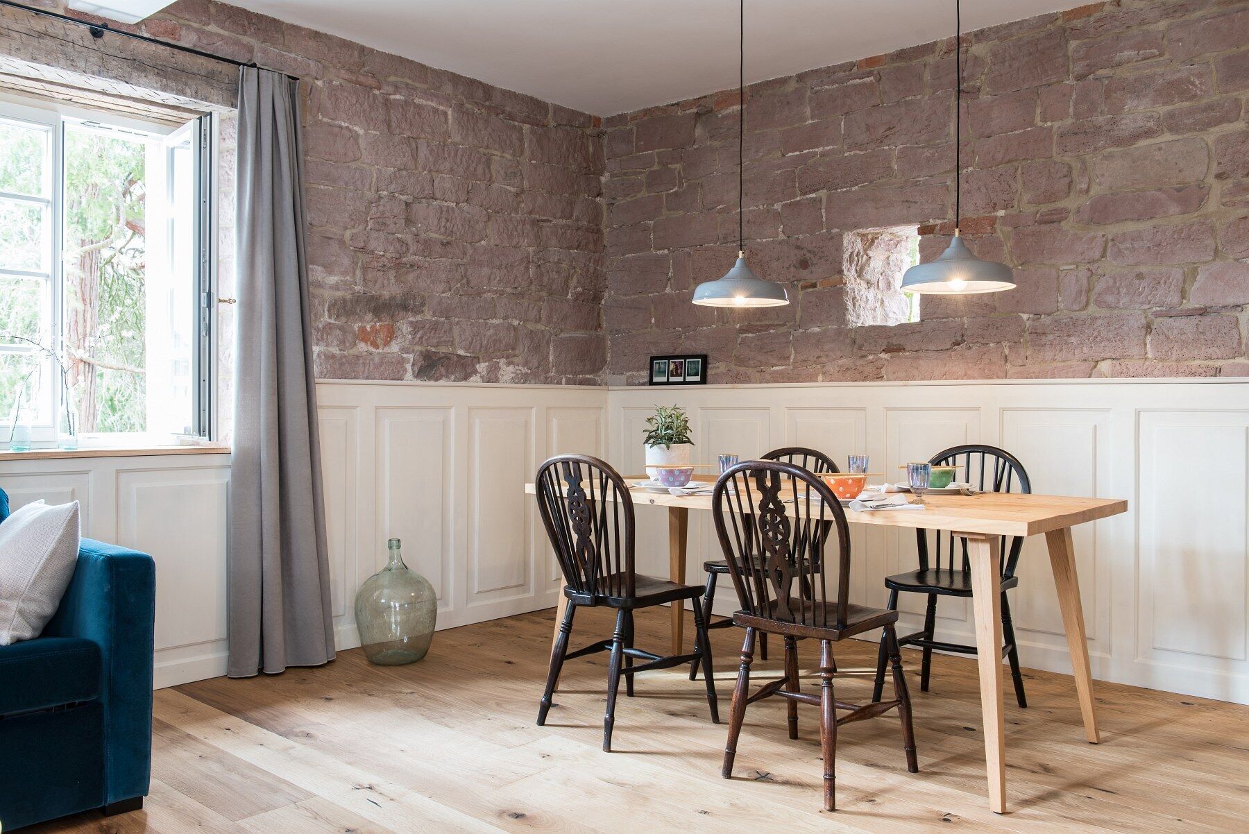 A dining area with a wooden table, four chairs, two hanging pendant lights, and a brick wall.