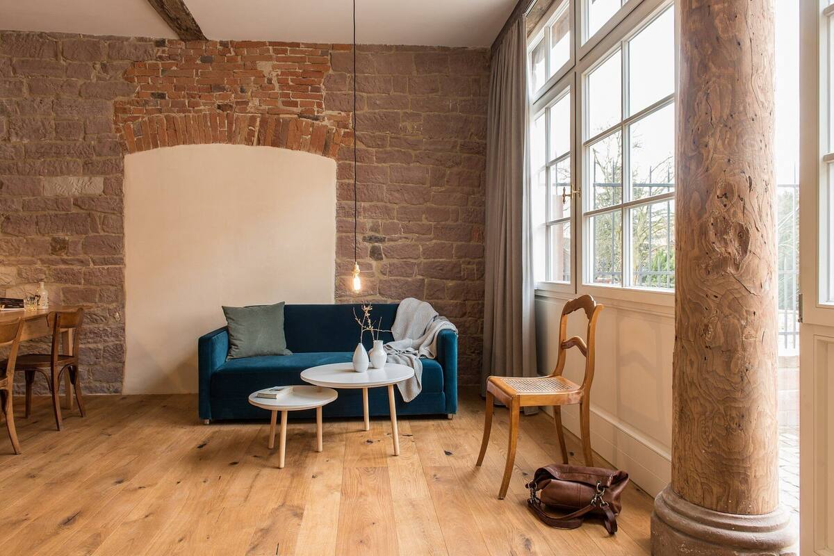Une pièce chaleureuse avec un mur en briques, un canapé bleu, deux petites tables, une chaise en bois et une grande fenêtre.