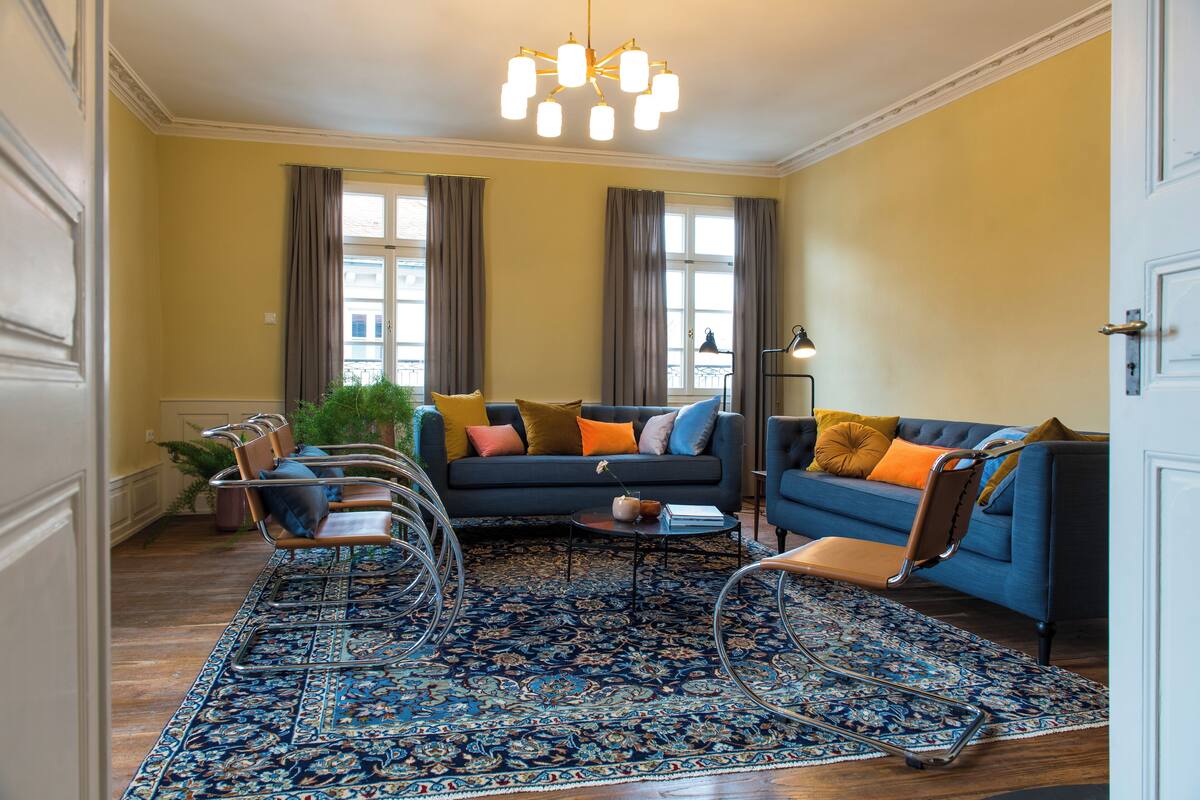 Un salon avec un canapé bleu, des fauteuils en cuir marron, un tapis à motifs et un lustre.